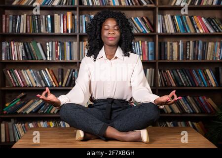 Donna d'affari afro-americana concentrata con capelli ricci medita seduta in yoga ponga il loto sulla parte superiore di legno marrone della tabella alle librerie dell'ufficio Foto Stock