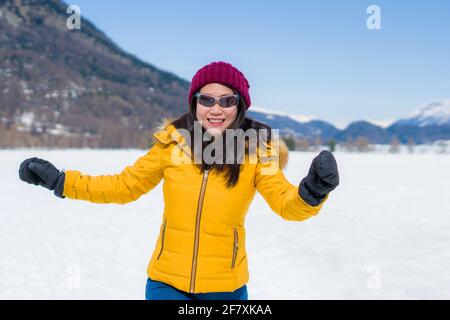 Giovane donna cinese asiatica felice e allegra in giacca invernale e cappello che corre divertente su lago ghiacciato coperto di neve Sulle Alpi svizzere godendo Foto Stock