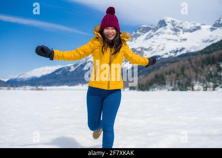 Giovane donna cinese asiatica felice e allegra in giacca invernale e cappello che corre divertente su lago ghiacciato coperto di neve Sulle Alpi svizzere godendo Foto Stock