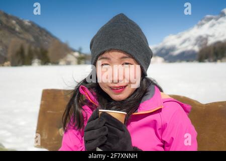 Gelo vacanze invernali - giovani felici e bei cinesi asiatici donna in panchina al lago ghiacciato e neve montagne bere caffè o cioccolata calda e.