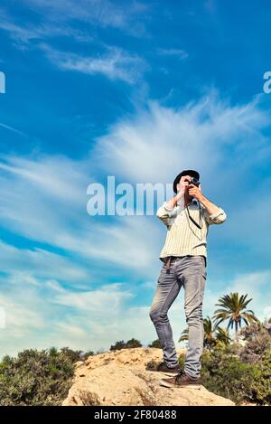 Un turista avventuroso scatta foto con la sua fotocamera d'epoca una zona con palme e un bel cielo dentro lo sfondo Foto Stock
