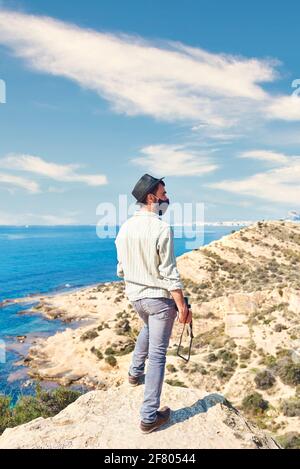 Un giovane tiene la sua macchina fotografica mentre ha una costa con il mare sullo sfondo, è mattina presto e la foto è molto luminosa Foto Stock