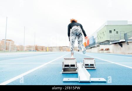 Vista posteriore di giovane forte sportivo che comincia a correre veloce dai blocchi di partenza durante la pista e l'allenamento sul campo allo stadio Foto Stock