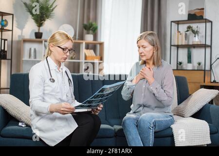 Donna anziana preoccupata e disturbata, che tiene le mani sul cuore, ascolta il medico femminile, la visita a casa e spiega la scansione a raggi X. Donna dottore consulta il suo paziente a casa. Foto Stock