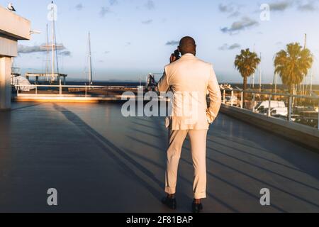 Vista da dietro di un ragazzo nero in un abito elegante che parla al telefono mentre si sta in piedi su una zona costiera tramonto; uomo calvo africano in un costume bianco Foto Stock
