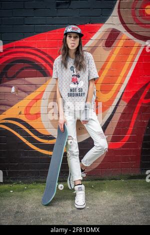 Ragazza adolescente con skateboard che si stacca la lingua fuori e si alza vicino al muro urbano con graffiti colorati a Shorditch a Londra , Regno Unito . Foto Stock