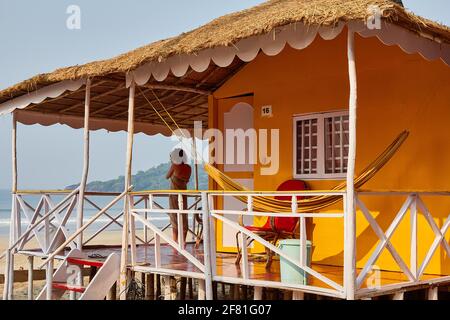 Donna turistica in piedi sul portico di una capanna gialla spiaggia con tetto di paglia e un amaca gialla appesa al tetto a Palolem Beach, Goa Foto Stock