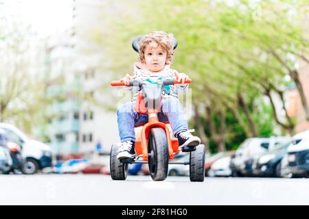 Ragazzo di due anni e mezzo seduto su di lui triciclo sulla strada in mezzo alla strada Foto Stock