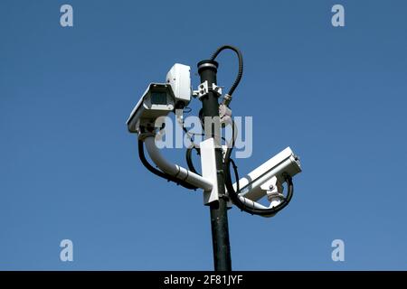 Due videocamere di sorveglianza bianche e altri accessori di sicurezza assortiti sulla parte superiore di un palo alto, contro un cielo blu chiaro. Ottawa, Ontario, Canada. Foto Stock