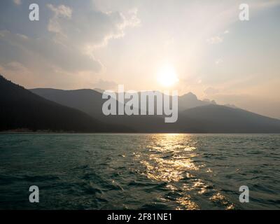 tramonto dietro un'alta montagna in estate con acqua di un lago in primo piano e fumo nel aria Foto Stock