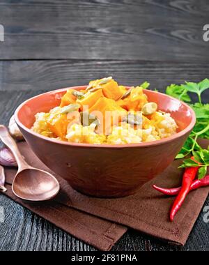 Porridge di miglio con salsa piccante di zucca e semi in un recipiente di argilla su un tovagliolo, pepe caldo, radice di zenzero, aglio e prezzemolo su sfondo di legno Foto Stock