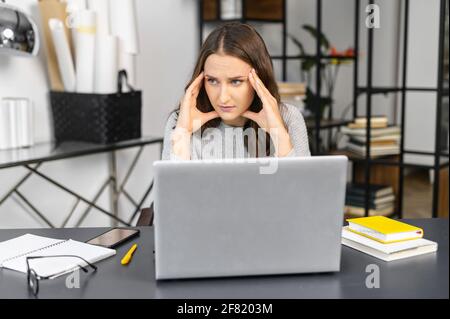 Donna preoccupata si siede alla scrivania di fronte al computer portatile in un ufficio moderno, guarda via e tiene le tempie, femmina in abbigliamento casual sente un mal di testa, preoccupato di risolvere le attività Foto Stock