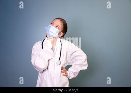 bella ragazza giovane con 2 trecce vestita da medico con maschera protettiva e stetoscopio davanti a sfondo blu Foto Stock