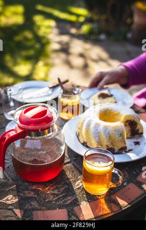 Tè in tazza e teiera con torta in padella sul tavolo. Serve dessert e bevande calde nel giardino di primavera Foto Stock