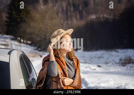 Giovane donna con cappello che si scalda in coperta sulla luce del sole e che riposa accanto all'auto durante il viaggio invernale su strada. Godimento di una giornata di sole in inverno Foto Stock