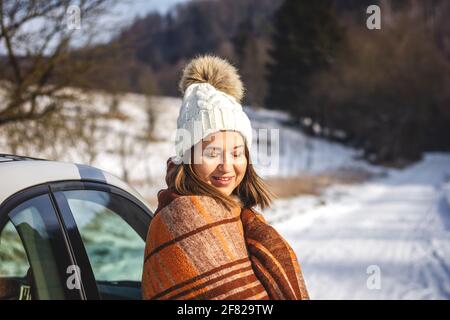 Giovane donna si scalda in coperta sulla luce del sole e riposa accanto alla macchina durante la vacanza invernale. Donna caucasica sorridente con gli occhi chiusi Foto Stock