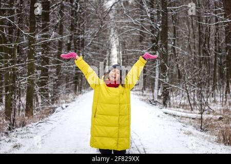 Felice giovane donna godendo di passeggiata invernale all'aperto nella foresta. Donna che indossa cappotto giallo e cappello a maglia in natura innevata Foto Stock