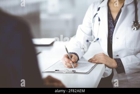 Donna-medico sconosciuto sta scrivendo alcune raccomandazioni mediche al suo paziente, mentre stanno sedendo insieme alla scrivania nel gabinetto in una clinica Foto Stock