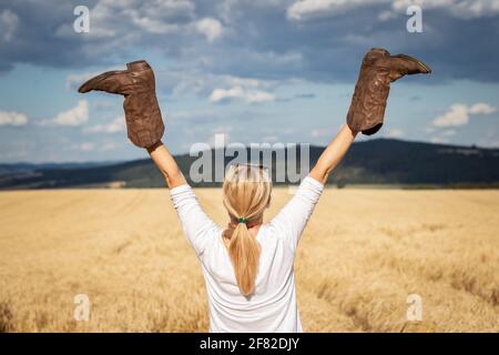 Donna felice che tiene gli stivali da cowboy in mano sollevati all'aperto. Cowgirl godendo estate in campagna. Sensazione di libertà Foto Stock