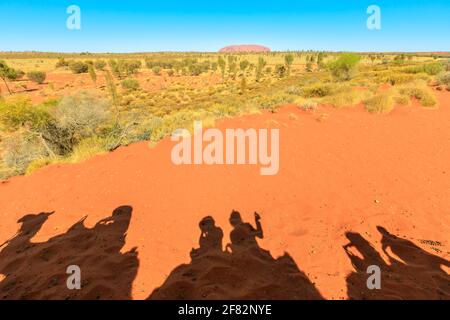 Uluru, Australia - Agosto 2019: L'ombra della gente di cavalcare il cammello nel deserto australiano del territorio del Nord con la roccia di Uluru Ayers. Attività popolare a. Foto Stock