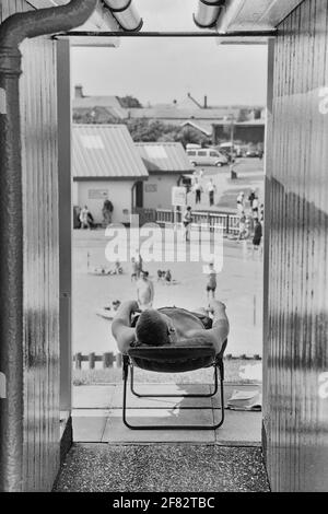 Un uomo che prende il sole accanto al suo chalet sulla spiaggia presso la piscina a spruzzo del Queen's Park. Mablethorpe. Lincolnshire. Inghilterra, Regno Unito Foto Stock