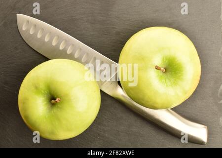 Due mele biologiche, succose, mature, dolci, verdi con coltello metallico, primo piano, sullo sfondo di un piatto di portata di ardesia. Foto Stock