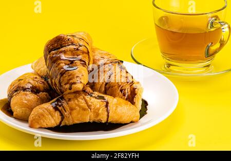 Ammira il mucchio di croissant fatti in casa con la salsa al cioccolato in un piatto di ceramica bianca e una tazza di tè caldo su sfondo giallo con percorso di ritaglio. Foto Stock