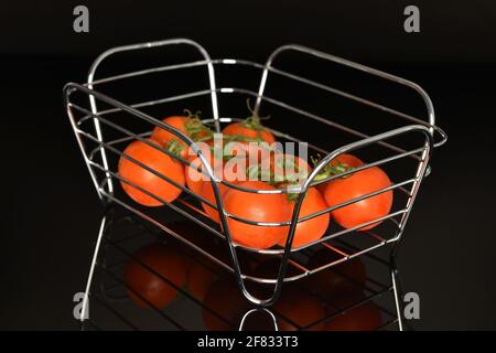 Un gruppo di diversi pomodori maturi, rossi brillanti, biologici, deliziosi cocktail, primo piano, con un cesto di filo metallico, su fondo nero. Foto Stock