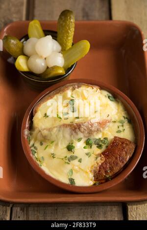 Un piatto fatto in casa di Raclette fuso, una patata al forno e salsiccia di chorizo alla griglia servita con cetriolini e cipolle sottaceto. Raclette ha avuto origine in Foto Stock