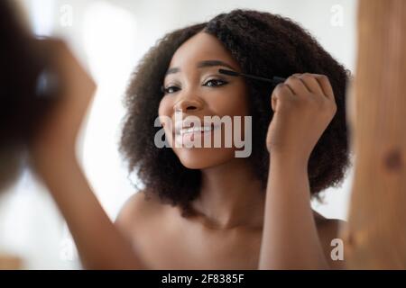 Splendida donna nera che applica mascara sulle sue ciglia di fronte di specchio a casa Foto Stock