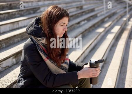 triste donna penosa che beve il tè dalla tazza termica sui gradini delle scale in strada. tempo di quarantena - caffè chiusi, cibo da asporto e bevande Foto Stock