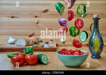 Gli ingredienti andalusi del gazpacho cadono nella ciotola con una mano che aggiunge il sale e insieme delle verdure sul lato sinistro, con il pane dietro, fuori fuoco. Foto Stock