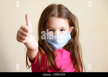 Capretto in maschera facciale per la protezione al virus corona mostra il pollice in su, ritratto della bambina graziosa che indossa la maschera medica a causa di COVID-19. Carino bambino stayin Foto Stock