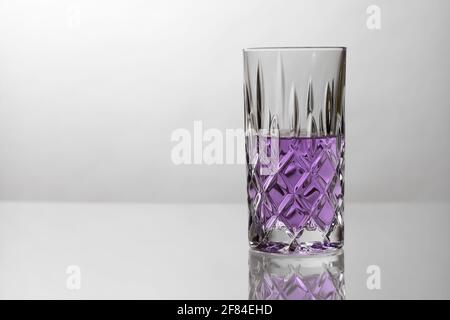 Bicchiere da cocktail mezzo completo con bevanda viola. Cristallo su una superficie riflettente. Spazio per il testo a sinistra. Foto Stock