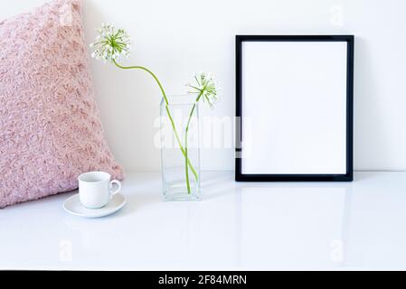 Mockup nero vuoto della cornice. Piccoli fiori freschi di aglio bianco (allium napolitanum) in vaso di vetro. Tazza di caffè, cuscino ricamato rosa su wh lucido Foto Stock