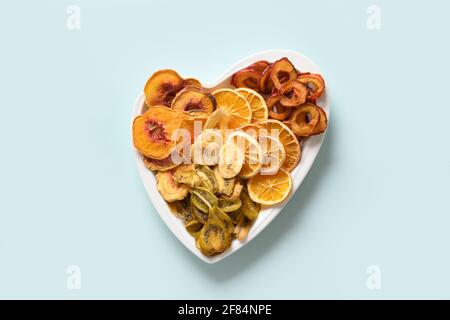 Prugne essiccate a fette, kiwi, patatine di frutta di pesca a forma di piatto di cuore su sfondo blu, essiccazione domestica. Snack vegan senza zucchero cibo. Vista dall'alto. Foto Stock