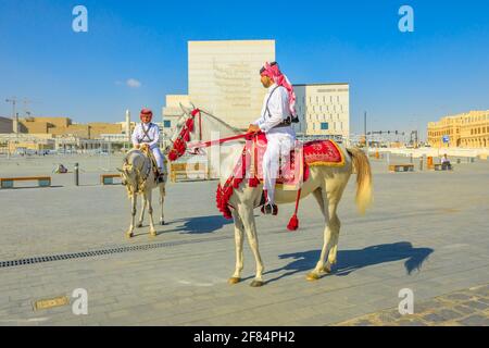 Doha, Qatar - 20 febbraio 2019: Due ufficiali di polizia di eredità in uniforme di Qatar tradizionale cavalcando cavalli arabi bianchi in piazza di Souq Waqif Foto Stock