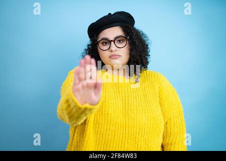 Giovane bella donna che indossa look francese con beretto e giallo maglione casual su sfondo blu isolato serio e facendo sosta canta con il palmo di Foto Stock