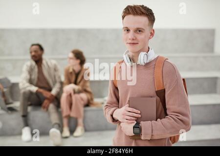 Vita ritratto di giovane studente di sesso maschile con zaino sorridente alla fotocamera mentre si trova in piedi nella moderna sala universitaria, copia spazio Foto Stock