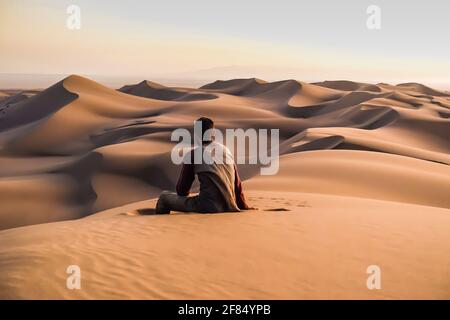 Una vista posteriore di meditativo solo giovane seduto sopra la sabbia nel deserto guardando le dune di sabbia Foto Stock