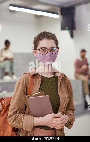 Verticale vita in su ritratto di giovane studentessa indossando maschera e guardando la macchina fotografica mentre si è in piedi nella moderna sala della scuola Foto Stock