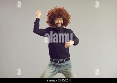 Ragazzo felice con pazzo divertente capelli ricci danzare stile gangnam su sfondo grigio studio Foto Stock