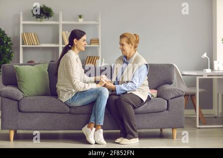 Figlia giovane cresciuta e madre anziana che tiene le mani seduta sul divano di casa Foto Stock