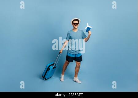 A lunghezza intera, il gioioso viaggiatore maschile caucasico in un cappello e occhiali da sole, vestito in estate, tiene una valigia blu, passaporto, biglietto e piccolo aereo, guarda la macchina fotografica, sorriso, sfondo blu isolato Foto Stock