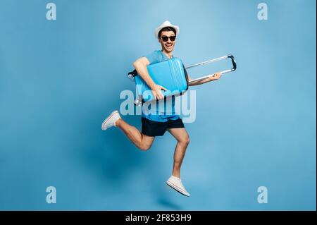 Foto a tutta lunghezza di un uomo caucasico felice stupito in un occhiali da sole e cappello, in abbigliamento casual, saltando con una valigia in mano su uno sfondo blu. Eccitato per una vacanza attesa da tempo, sorridente Foto Stock