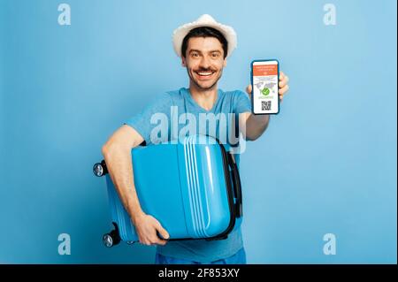 Certificato elettronico di vaccinazione dell'infezione da COVID19. Il ragazzo felice defocused tiene smartphone con certificato elettronico di vaccinazione e valigia in mani, stare su sfondo blu isolato Foto Stock