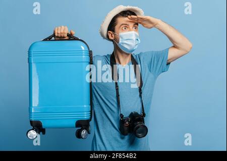 Gioioso viaggiatore in un cappello e maschera medica, vestito in t-shirt blu, contiene una valigia blu, con fotocamera, guarda in lontananza, felice di una vacanza attesa da tempo, isolato sfondo blu Foto Stock