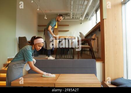 Vista laterale ritratto di due tavoli per la pulizia di giovani camerieri in caffetteria con caldi accenti di legno, spazio per la copia Foto Stock