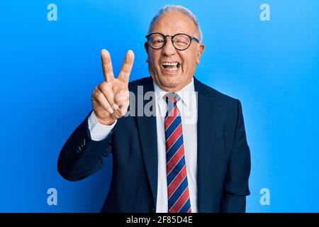 Uomo caucasico senior che indossa il costume da lavoro e legare sorridendo con felice faccia che si inarricia alla macchina fotografica facendo segno di vittoria con le dita. Numero due. Foto Stock