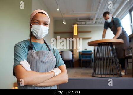 Vita in su ritratto della cameriera giovane che indossa la maschera e grembiule guardando la macchina fotografica mentre si trova in una caffetteria con uomo tabelle di pulizia in background, covid sicuro Foto Stock